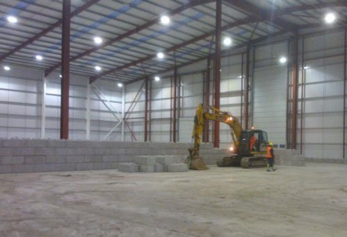 Excavator installing a low interlocking block wall inside an empty steel-framed warehouse