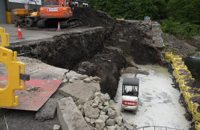 Excavated trench with concrete block walling and an excavator on a roadside site